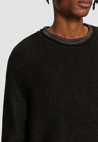 Schwarzer strukturierter Strickpullover mit lockerer Passform und weitem Ausschnitt, über einem dunklen, gerippten Unterhemd getragen.