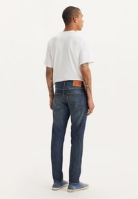 Μπλε τζιν παντελόνι με μεσαίο ξεβάμμα, ετικέτα πίσω και διακριτικές λεπτομέρειες ραφής, σε συνδυασμό με λευκό κοντομάνικο μπλουζάκι και μπλε αθλητικά παπούτσια.