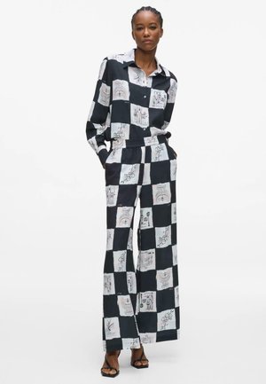 Femme debout les mains dans les poches, portant une chemise à carreaux noir et blanc et un pantalon à jambes larges avec des dessins abstraits, sandales noires.