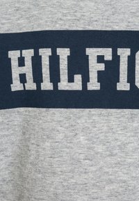 Harmaa puuvillainen t-paita, jossa on tummansiniharmaa vaakasuora raita ja kirkasvalkoinen teksti "HILFIGER" rinnassa.