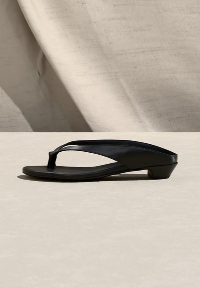 Μαύρο σανδάλι με χαμηλό τακούνι και σχεδίαση με λουράκι ανάμεσα στα δάχτυλα, με λεία επιφάνεια και ελαφρώς καμπυλωτή σόλα πάνω σε ουδέτερη επιφάνεια και υφασμάτινο φόντο.