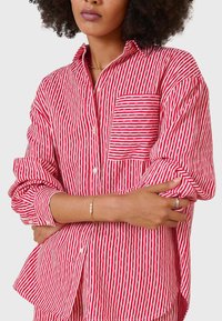 Red and white striped button-up shirt with a chest pocket, long sleeves, and soft fabric texture. Accents include rolled sleeves and curved hem.