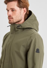 Olive green waterproof jacket with a high collar, adjustable hood, and a logo patch on the left shoulder. Smooth texture, modern design.