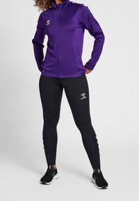 Fioletowa sportowa kurtka na zamek z wysokim kołnierzem, w zestawie z czarnymi dopasowanymi legginsami z logo. Całość dopełniają czarne buty.