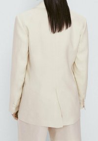 Beige maßgeschneiderter Blazer mit einer strukturierten Silhouette, einem einzelnen Rückenschlitz und geknöpften Manschetten. Der Stoff wirkt glatt mit einer dezenten Textur.