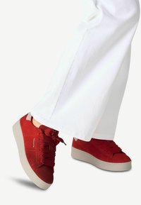 Sneaker in suede rosso con punta tonda, suola spessa grigia e lacci testurizzati, abbinati a pantaloni bianchi a gamba larga. Dettagli di cucitura notevoli visibili.