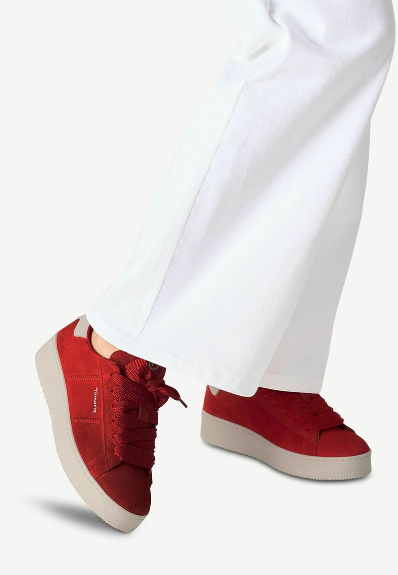 Sneaker in suede rosso con punta tonda, suola spessa grigia e lacci testurizzati, abbinati a pantaloni bianchi a gamba larga. Dettagli di cucitura notevoli visibili.