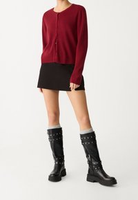 Ruby red ribbed cardigan, black mini skirt, and knee-high black boots with silver studs and grey knit cuffs. Flat sole, chunky design.