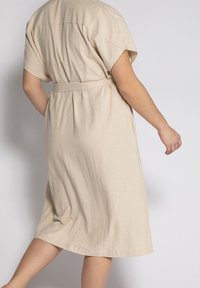 Abito in lino beige con maniche corte, vita cintrata, orlo leggermente svasato e texture sottile, che mostra un design semplice e minimalista.
