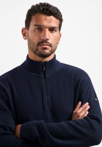 Man met kort krullend haar en een baard, gekleed in een marineblauwe, gestructureerde trui met een kwart rits, met gekruiste armen, die recht vooruit kijkt.