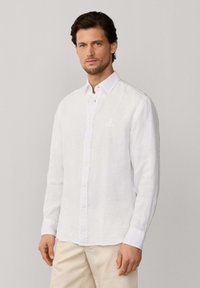Homme portant une chemise blanche à manches longues boutonnée et un pantalon beige, debout devant un fond gris clair uni.