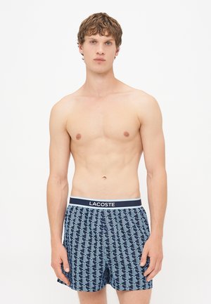 Boxers pour hommes en bleu marine avec un motif géométrique en bleu clair. Présentent une ceinture contrastante avec le logo "LACOSTE" et une texture de tissu douce.