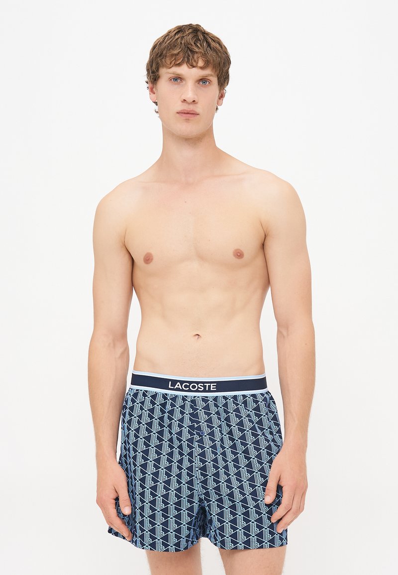Boxeri pentru bărbați de culoare albastru marin cu un model geometric în albastru deschis. Dispune de un brâu contrastant cu logo-ul "LACOSTE" și o textură moale a materialului.
