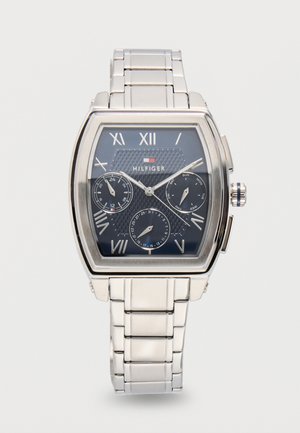 Sølvfarget metallarmbåndsur med svart urskive, romertall, tre underskiver for 24 timer, dag og dato, og Tommy Hilfiger-logo.