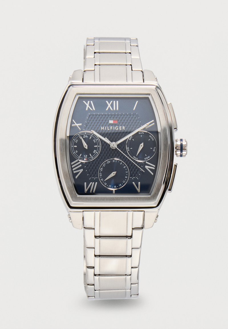 Ασημένιο μεταλλικό ρολόι χειρός με μαύρο καντράν, ρωμαϊκούς αριθμούς, τρεις υποκαντράν για 24ώρο, ημέρα και ημερομηνία, και λογότυπο Tommy Hilfiger.