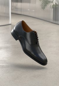 Scarpa da uomo in pelle nera con una texture liscia, caratterizzata da un design minimalista con lacci e una punta tonda, su uno sfondo neutro.