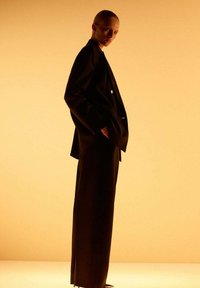 Persoon met slicked-back haar in een losse zwarte suit met handen in de zakken op een warme gele achtergrond.