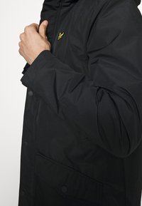 Czarna pikowana kurtka o gładkiej fakturze, z zamkiem błyskawicznym i zatrzaskami. Na piersi widoczne żółte logo oraz skośna przednia kieszeń.
