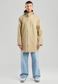 Beige waterproof coat with a high collar, front snap closures, side pockets, and a hood, paired with light blue wide-leg jeans.