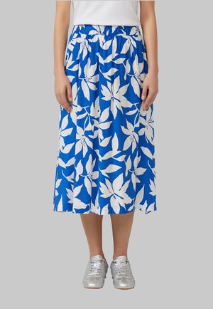 Une femme porte une jupe midi bleue à grands motifs floraux blancs, associée à des baskets argentées et un haut blanc, debout devant un fond gris.
