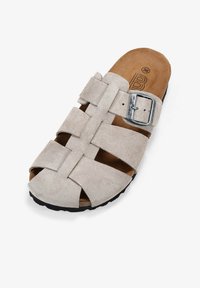 Sandalia deslizante de ante en gris claro con diseño de tiras cruzadas, hebilla metálica ajustable y plantilla contorneada marrón sobre una suela negra texturizada.