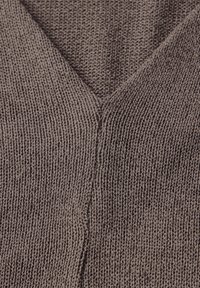 Ruskea neulottu kangas, jossa on V-kaula-aukko. Siinä on teksturoitu kudonta pystysuuntaisilla ribbineuleilla ja näkyvillä saumoilla.