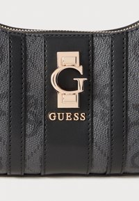 Juodas oda rankinės su vertikaliomis struktūrinėmis juostelėmis. Papuošta auksinės spalvos „G“ logotipu ir žodžiu „GUESS“ auksiniu tonu ant poliruoto metalo plokštelės.