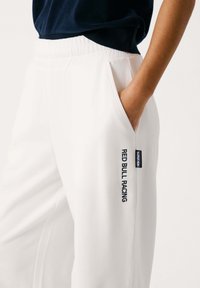 Baltos sportinės kelnės su elastinga juosta, šoniniais kišenėmis ir tamsiai mėlynu "RED BULL RACING" logotipu kairėje pusėje. Minkšta medžiagos tekstūra.