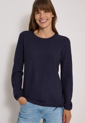 Femme souriante, portant un haut texturé bleu foncé à manches longues et un jean bleu clair, debout devant un fond clair uni.