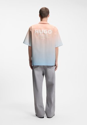 Άτομο που βλέπει προς τα πίσω φοράει κοντομάνικο με διαβάθμιση από πορτοκαλί σε μπλε και λευκό κείμενο "HUGO", συνδυασμένο με φαρδιά γκρι παντελόνια.