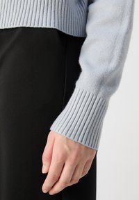 Main reposant sur le côté, vêtue d'une manche de pull en maille côtelée gris clair et d'un pantalon noir sur un fond uni.