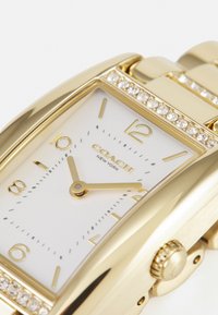 Montre en métal doré avec un boîtier rectangulaire, un cadran blanc, des index horaires dorés et des accents en cristal autour de la lunette. Design simple et élégant.