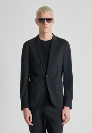 Giovane uomo con capelli biondi corti che indossa occhiali da sole scuri oversize, blazer nero e pantaloni neri, in piedi contro uno sfondo semplice.