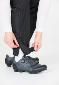 Sorte cykelbukser med sidelogoer og velcro, parret med mørkegrå sko med et kabelfastsystem og gummisåler.