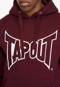 Bordo duks od pamučne mješavine, s velikim bijelim logotipom "TAPOUT". Ima prednji džep i kapuljaču sa vezicama.