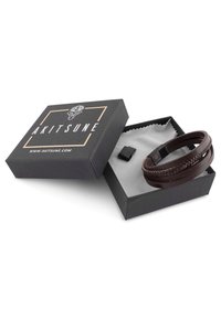 Pulsera de cuero marrón con detalle trenzado, presentada en una caja de regalo negra con un interior blanco, que presenta el nombre de la marca "AKITSUNE" en la tapa.