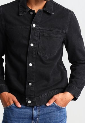 Mand iført en sort denimjakke med metalknapper, venstre brystlomme og blå jeans, hænderne i lommerne mod en hvid baggrund.