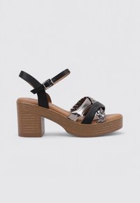 Sandalias de tacón alto en negro y plata metálica con un diseño frontal trenzado y un tacón cuadrado texturizado en marrón. Cuenta con una correa ajustable en el tobillo.