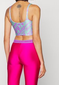 Sport-bh i ljus teal med lila grafisk text, med en kort design. Modellen bär glansiga rosa leggings med varumärkesmidja.