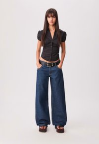Zwarte knoopblouse met pofmouwen, gecombineerd met een wijd uitlopend blauw denim broek en een zwarte riem; model draagt bruine sandalen.