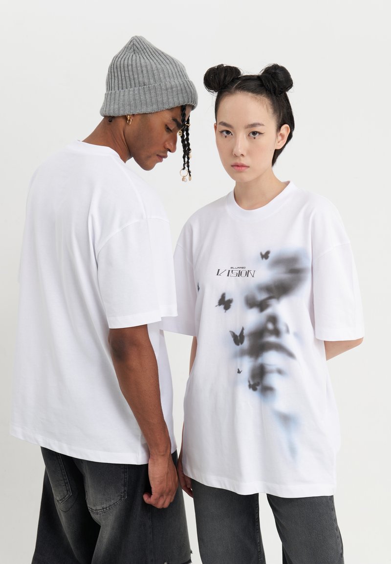 Camisetas oversized blancas de algodón, con un gráfico de mariposa. Un modelo lleva un gorro de punto gris; el otro tiene el cabello recogido en moños.