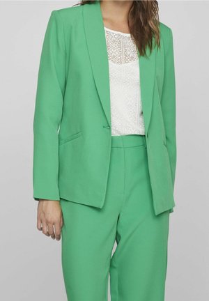 Femme portant un costume tailleur vert vif avec un chemisier blanc en dentelle, debout devant un fond gris clair uni.