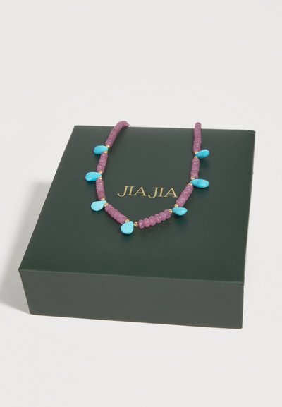 Κολιέ με μωβ χάντρες και μπλε πέτρες σε σχήμα σταγόνας, εμφανίζεται σε μια σκούρα πράσινη θήκη με χρυσό κείμενο "JIA JIA".