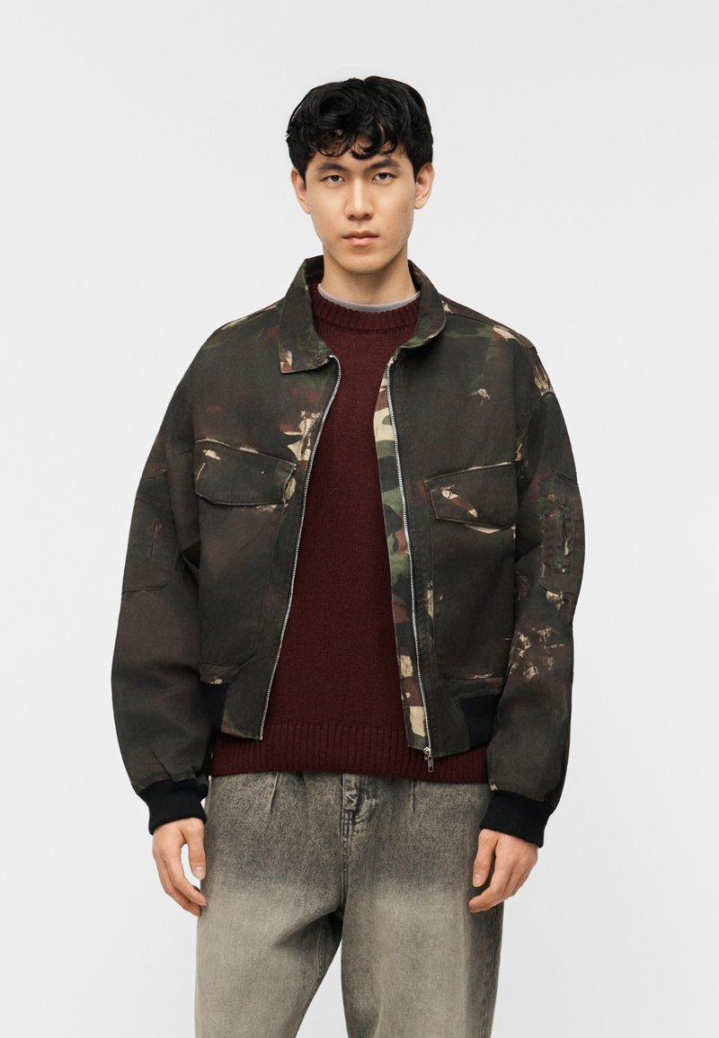 Νεαρός άνδρας που φοράει σκουρόχρωμο μπουφάν με φερμουάρ σε καμουφλάζ πάνω από μπορντό πουλόβερ και ξεθωριασμένο γκρι τζιν, στέκεται μπροστά σε απλό φωτεινό φόντο.