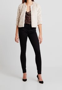 Chaqueta de un beige claro y textura, con un cuello tipo solapa, llevada sobre una blusa de estampado de leopardo y combinada con unos jeans skinny negros y tacones altos.