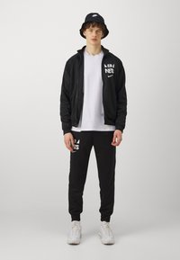 Giacca nera con zip e logo bianco, t-shirt bianca, pantaloni della tuta neri con grafiche, sneakers bianche e cappello a secchiello nero con logo.