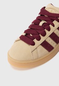 Bēšas zamšādas sporta kurpes ar bordo auduma šņorēm un trim bordo zamšādas svītrām sānos, novietotas uz gaiša fona.