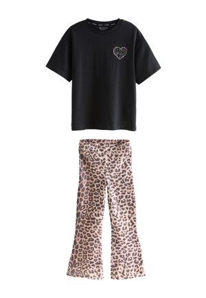 Młoda osoba z długimi włosami, ubrana w czarny luźny t-shirt z detalem serca oraz spodnie w panterkę, stoi na tle jednolitego jasnego tła.
