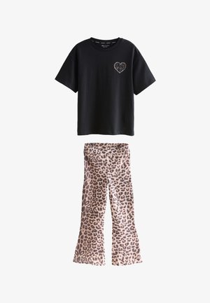 Młoda osoba z długimi włosami, ubrana w czarny luźny t-shirt z detalem serca oraz spodnie w panterkę, stoi na tle jednolitego jasnego tła.