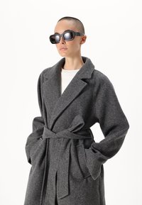 Sivina volnena jakna z pasom v pasu, šalom ovratnikom in velikimi žepi; kombinirana z ostrimi črnimi sončnimi očali, ki odražajo svetlobo.